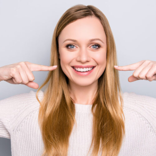 Portrait of joyful satisfied girl gesturing her beaming white healthy teeth with two forefingers looking at camera isolated on grey background. Orthodontic concept