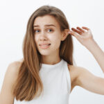 Stop lying my ears tired. Portrait of funny attractive woman with fair hair, pulling ear with hand and staring displeased at camera, feeling awkward while standing against gray background. Body language concept
